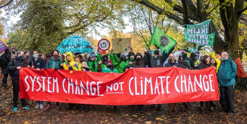 A large group of people gathered together outside holding a large banner which reads "system change not climate change"