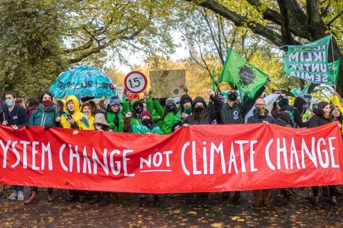 A large group of people gathered together outside holding a large banner which reads "system change not climate change"