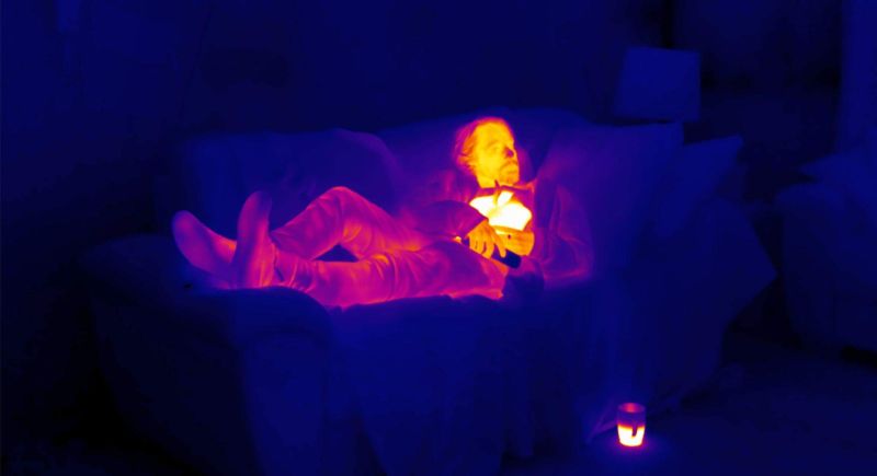 A man lies on a sofa holding a hot water bottle which glows as yellow on the thermal image.