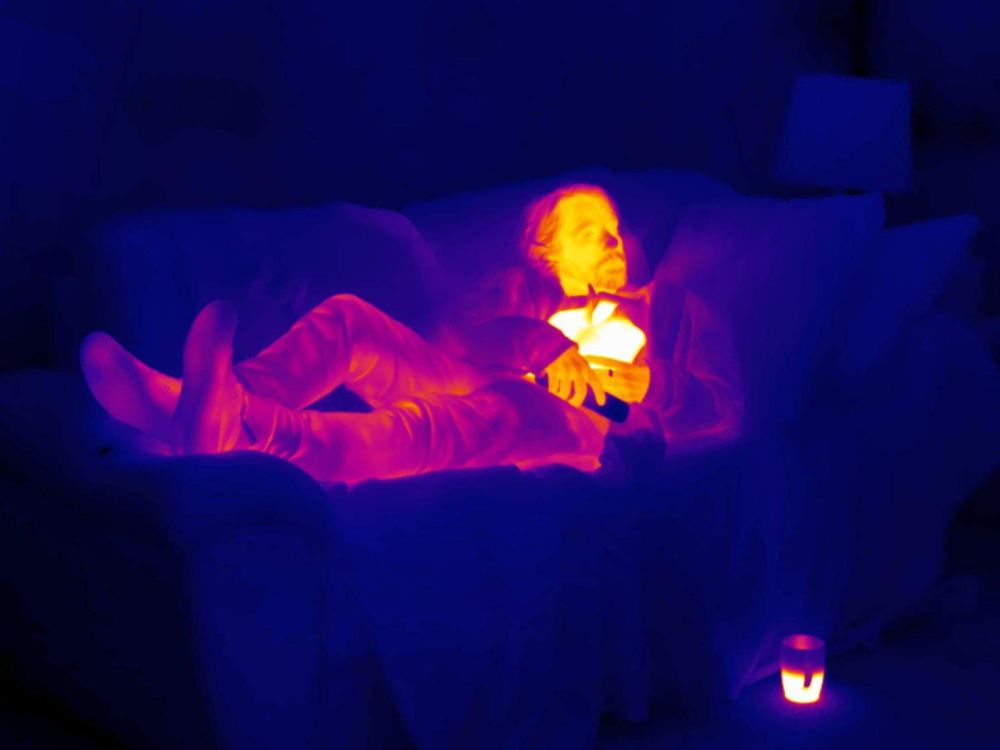 A man lies on a sofa holding a hot water bottle which glows as yellow on the thermal image.