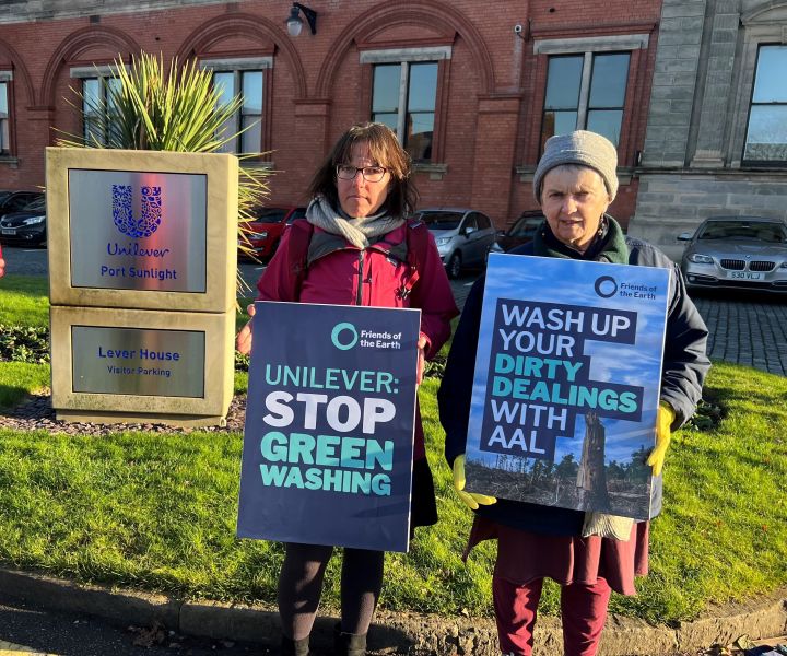 Two women stand in front of a building holding placards that read: "Unilever: stop greenwashing" and "Wash up your dirty dealings with AAL"
