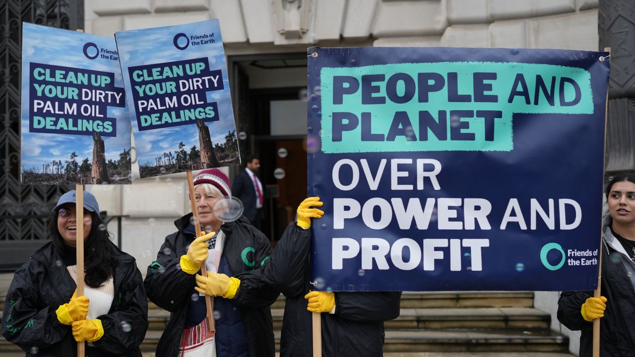 Campaigners stand outside a building holding placards and wearing yellow washing up gloves, while bubbles float in the air. The placards read: "People and planet over power and profit" and "Clean up your dirty palm oil dealings"