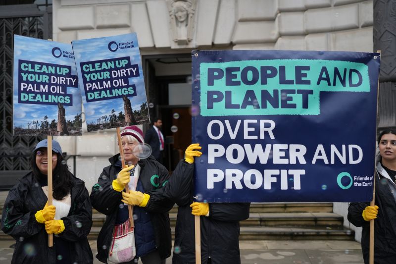 Campaigners stand outside a building holding placards and wearing yellow washing up gloves, while bubbles float in the air. The placards read: "People and planet over power and profit" and "Clean up your dirty palm oil dealings"