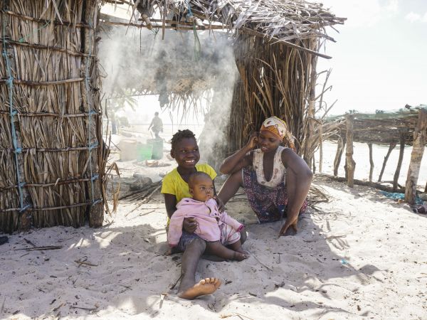 A young mother and her two children, one a baby, sitting on sand smiling. In the back are thatch homes on a beach