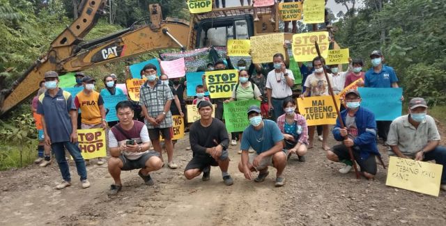 A group of villagers surround a digger holding signs saying "Stop the chop"