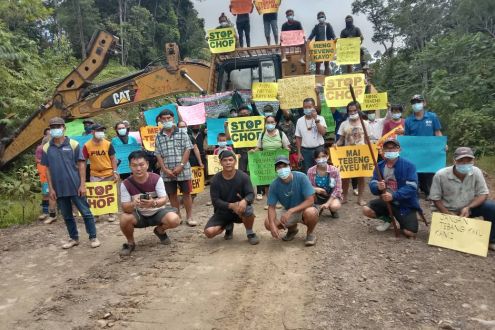 A group of villagers surround a digger holding signs saying "Stop the chop"