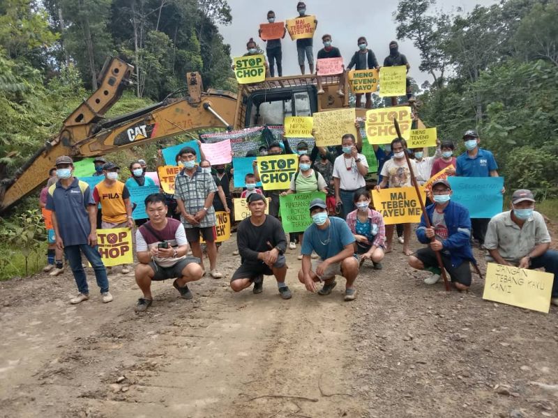A group of villagers surround a digger holding signs saying "Stop the chop"