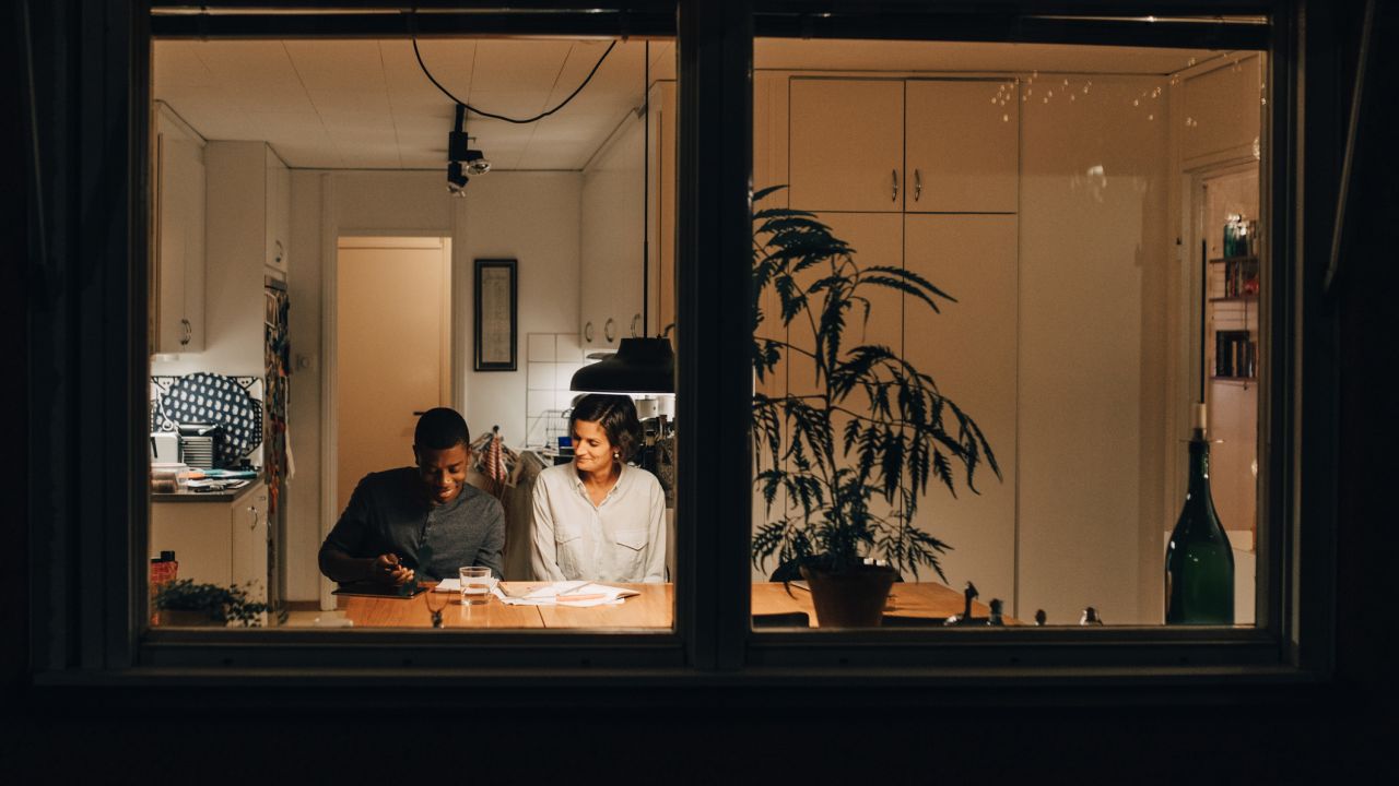 Looking through a dark window frame to a mother and son sat at a table lit with a hanging lamp
