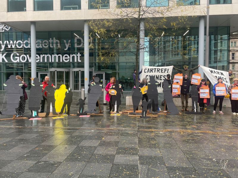 A group of people outside a government building holding black silhouette figures and placards demanding warm homes