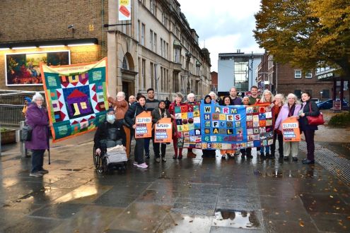 A group of people outside holding up their handmade quilt which reads "warm homes for all" alongside other placards demanding warm homes.
