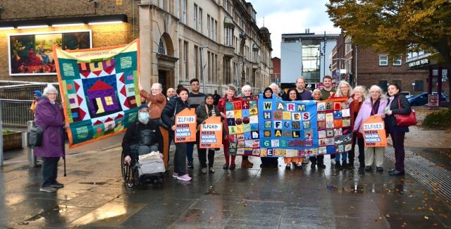 A group of people outside holding up their handmade quilt which reads "warm homes for all" alongside other placards demanding warm homes.