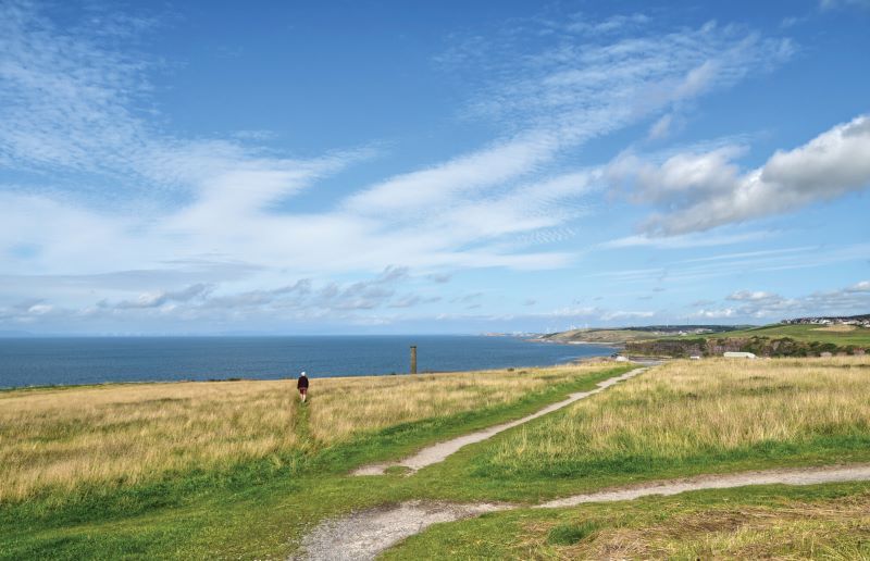 A man walking along a grassy cliff top path with the sea below and a wind farm on the horizon