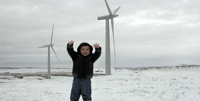 A little boy called Ben stands in front of 2 wind turbines at a Welsh community-owned wind farm