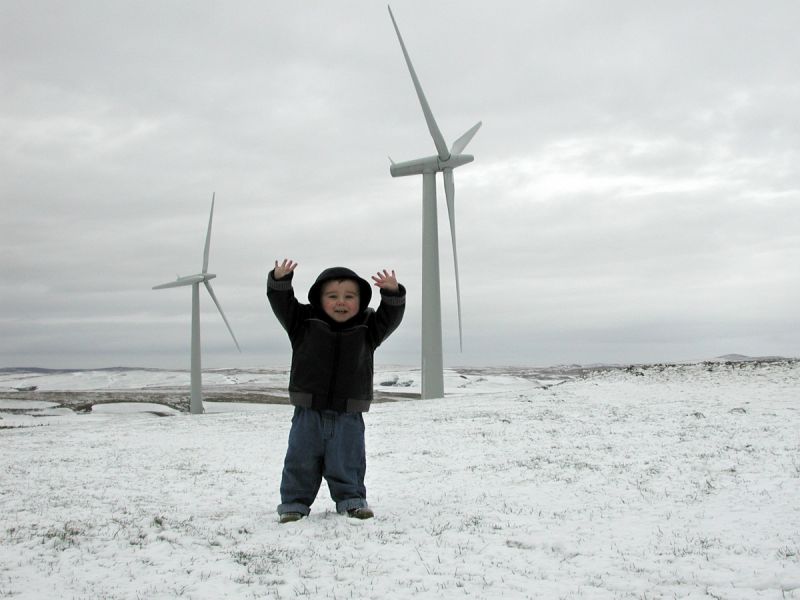 A little boy called Ben stands in front of 2 wind turbines at a Welsh community-owned wind farm