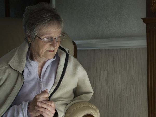 An older woman clutching her hands, wearing a coat indoors looking cold and sitting in the dark