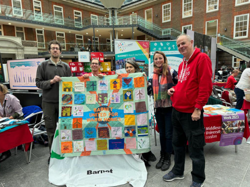 A group of 5 people proudly holding up a quilt in front of a stall with multiple squares reflecting their support for warm homes for all - including images of renewable energy sources. In the middle the quilt reads "Barnet united for warm homes".