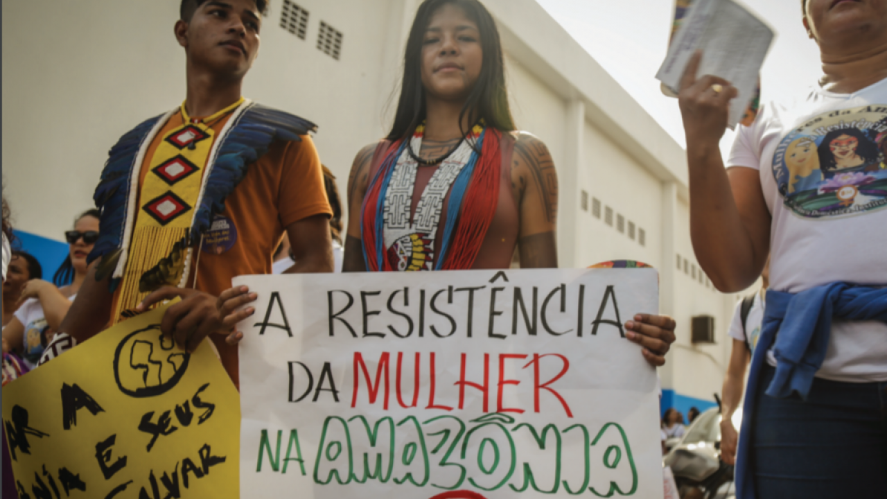 3 people protesting with one holding a sign which says 'A resistencia da mulher na amazonia' with a drawing of a symbol of women below