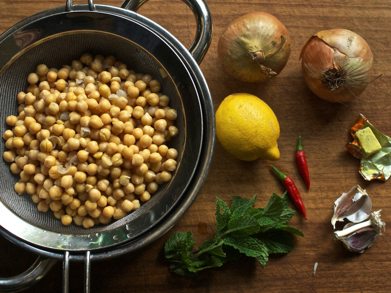 Chickpeas in sieve in bowl next to lemon, onions, chillies mint and stock cube