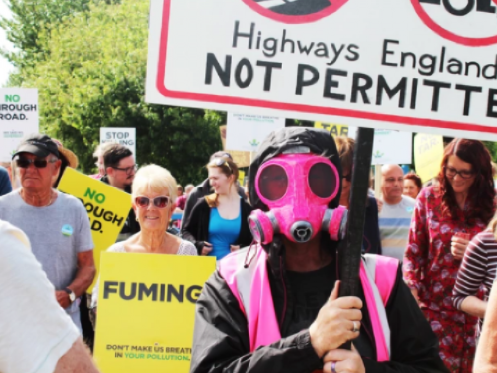 A protest with a person at the front wearing a pink gas mask and pink hi-vis holding a sign that says 'highways England, not permitted'