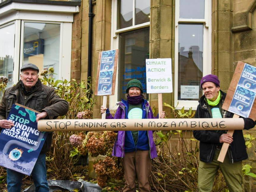 Three demonstrators protesting gas funding in Mozambique with a pipe and placards